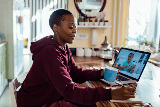 A woman taking notes while drinking coffee and watching a program on her laptop